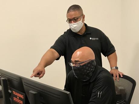 A man sits at a computer and receives help from another man who is standing behind him and pointing to the screen.